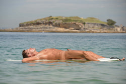 Ross Watson photograph of a naked man lying on surfboard in the ocean with an island in the background